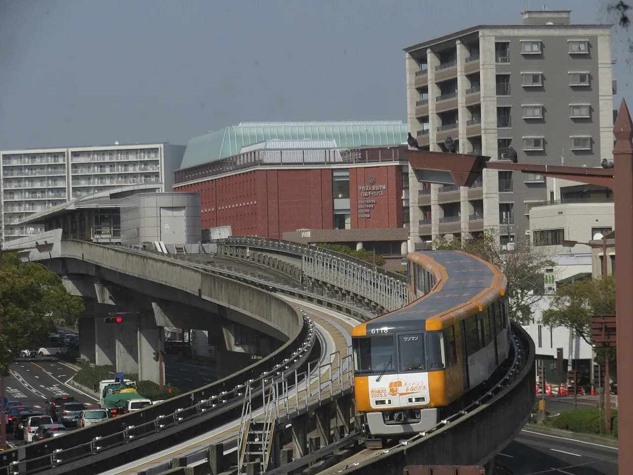 カーブする高架の軌道を、黄色い車体のアストラムラインの電車がこちらへ走ってくる。左に駅舎と車の並ぶ道路、奥にれんが色の校舎とマンションが建つ