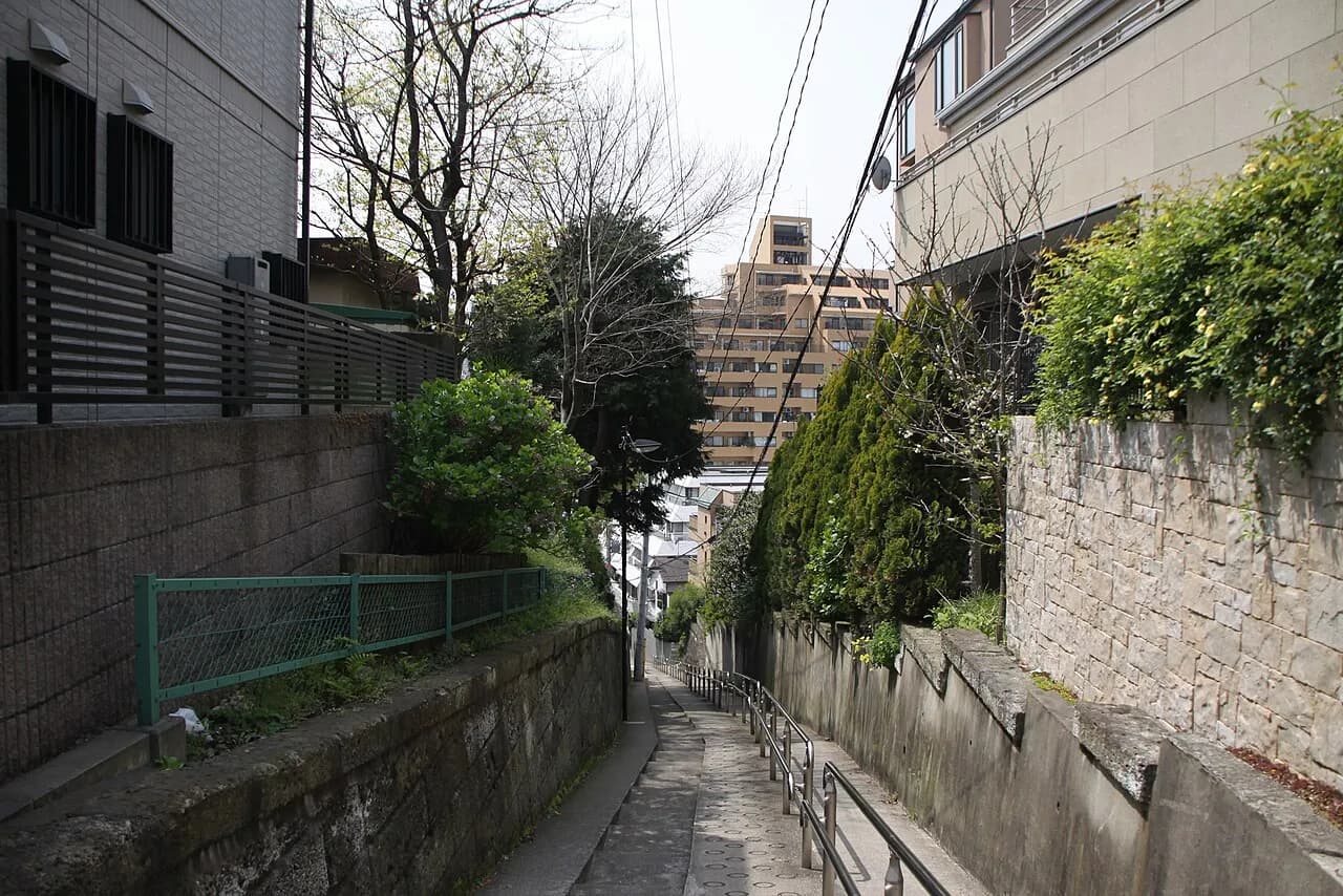石垣と植え込みに挟まれた細い下り坂の歩道。手すりが坂の下へ続き、両側に住宅とマンション、正面奥に中層のマンションが見える