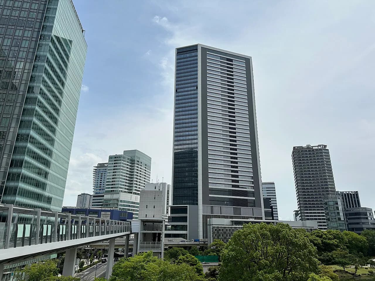 曇り空の下、浜松町駅東側の高層オフィスビル群。中央に縦長の東京ポートシティ竹芝オフィスタワー、左手前にゆりかもめの高架、手前に庭園の緑が広がる
