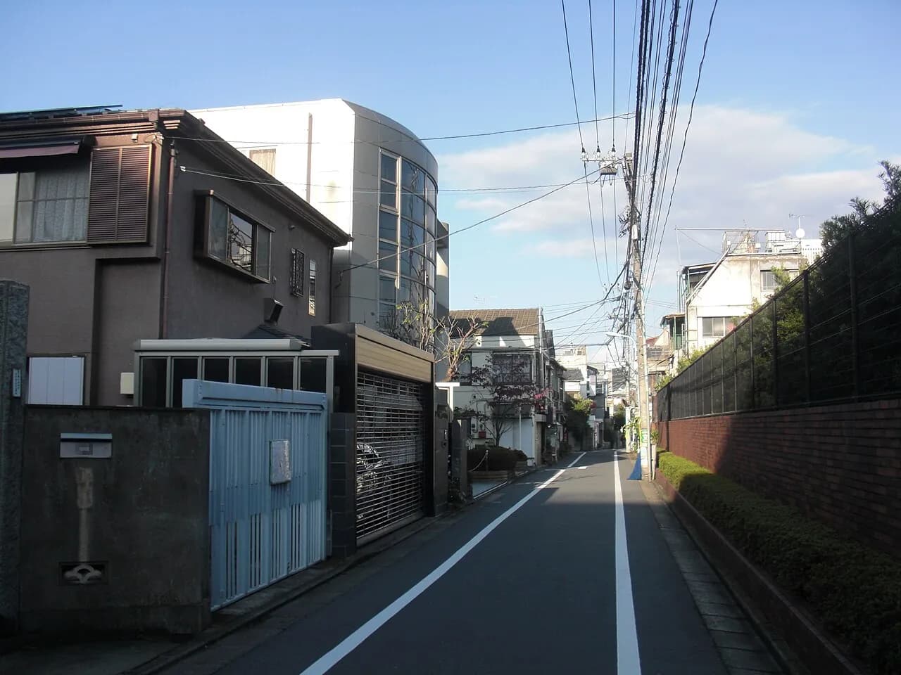 戸建て住宅と低層マンションに挟まれた静かな一方通行の道路。右側にれんがの塀と植え込みが続き、電線の向こうに晴れた空が見える