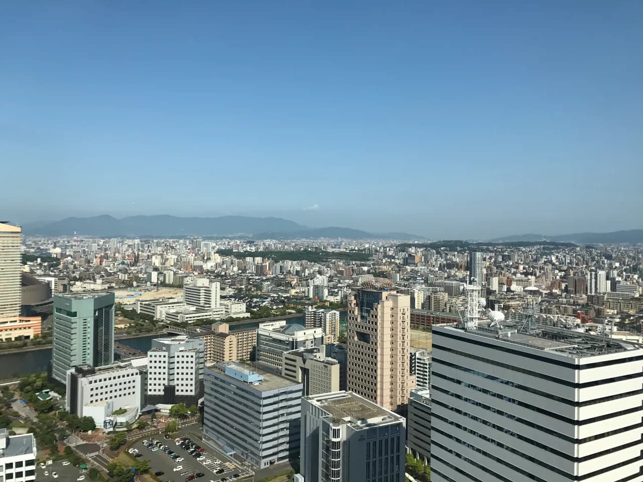 晴天の下、福岡タワーから見下ろした福岡市街。手前に百道浜のオフィスビル群と駐車場、川をはさんで中低層のマンションや住宅が奥まで続き、地平線に山並みが見える