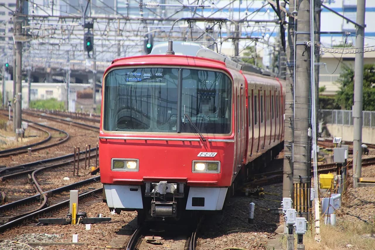 分岐する線路の上を、赤い名鉄の電車が正面からこちらへ走ってくる。背後に架線柱と高架橋、建物が見える