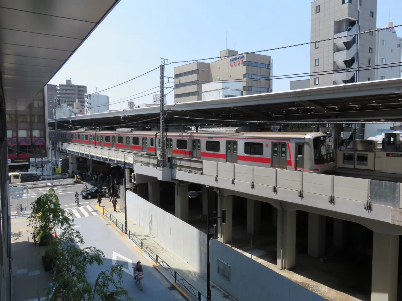 高い位置から見た中目黒駅の高架ホームと、停車中の赤帯の電車。高架の下には工事用の白い仮囲いがあり、左下に駅前の交差点と歩道が見える