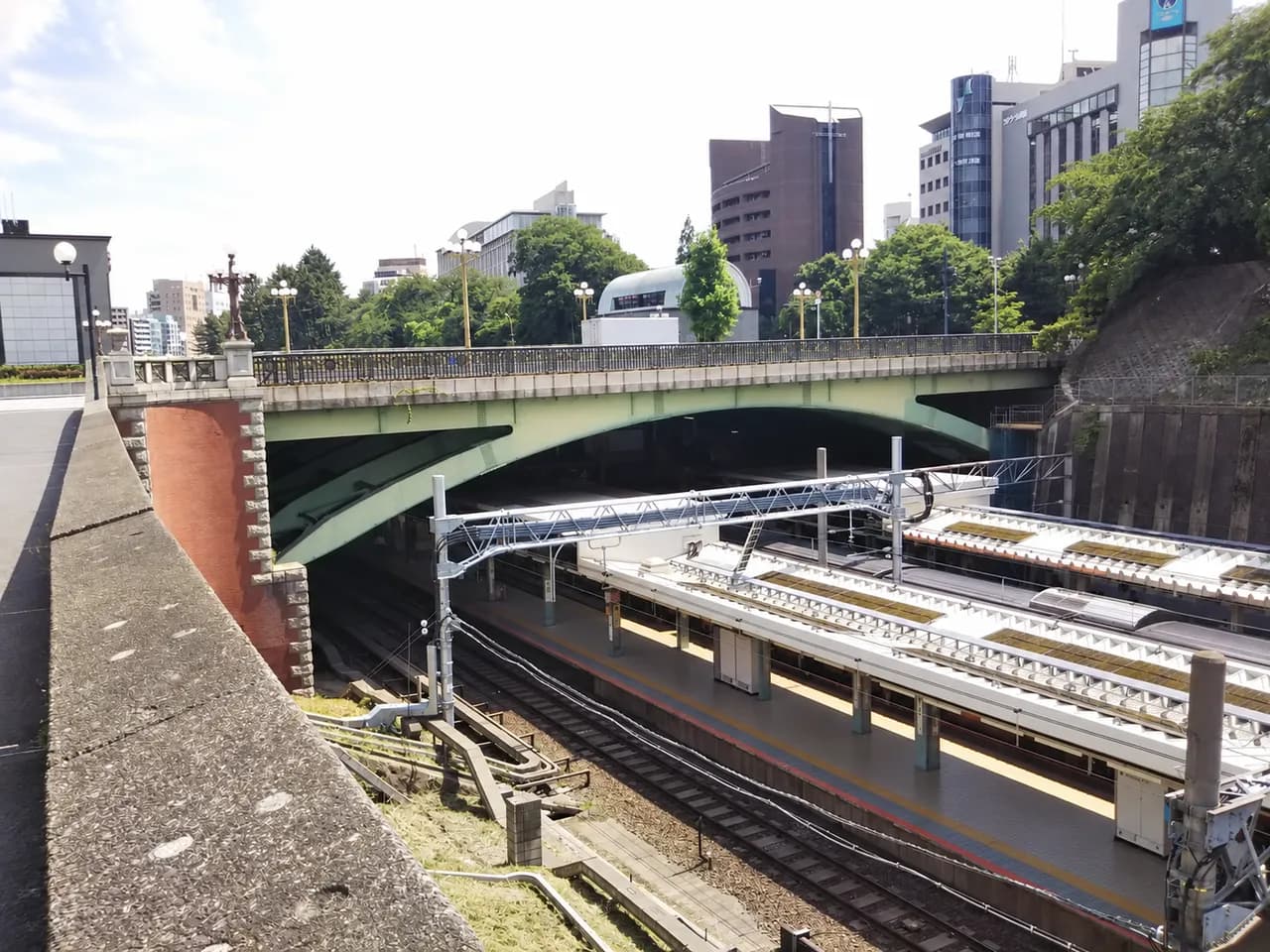 新宿通りの四谷見附橋を、その下のJR四ツ谷駅ホームと線路越しに見た眺め。緑色のアーチ橋の奥に街路樹とオフィスビルが並ぶ