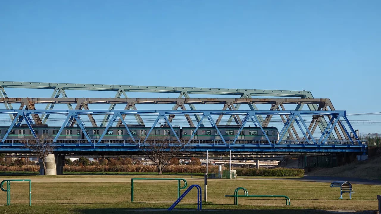 晴れた冬の午後、芝生の河川敷の奥に青い鉄橋が3本並び、千代田線の電車が渡っている。河川敷には運動器具やベンチがある