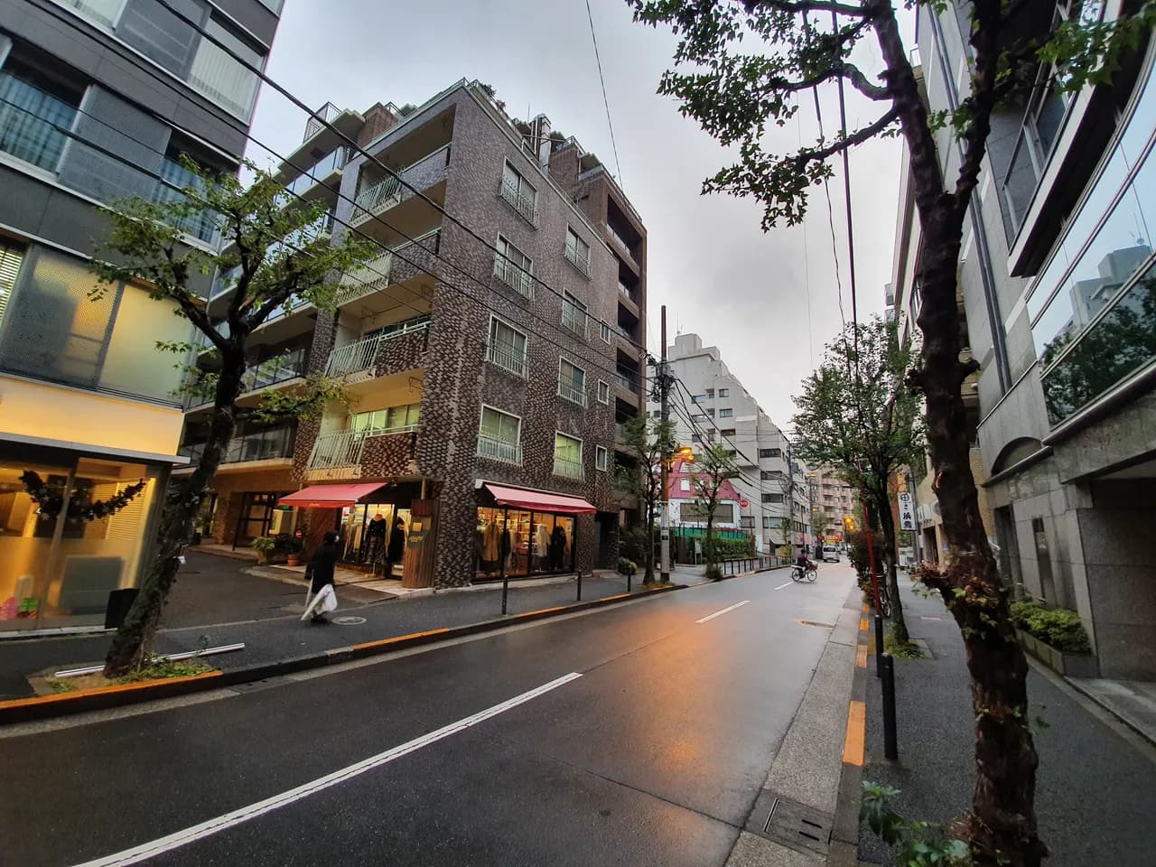 街路樹の並ぶ番町の通り。中央にれんが色タイルの中層マンションがあり、1階に赤い日よけの店舗。曇り空の夕方で路面がぬれている
