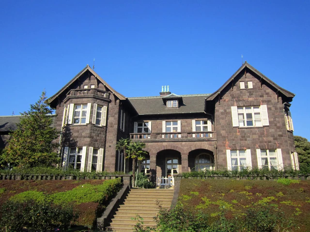青空の下に建つ旧古河庭園の石造りの洋館。正面の石段と植え込みの奥に、白い鎧戸の窓が並ぶ二つの切妻屋根の建物