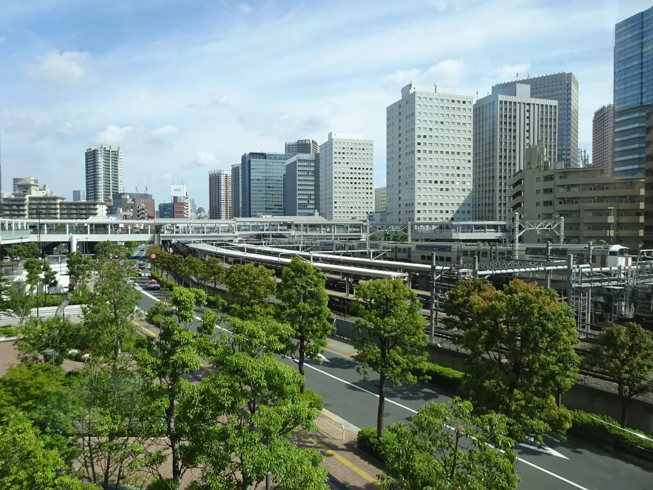 初夏の晴れた日に高い位置から見た大崎駅の周辺。手前に新緑の並木と線路沿いの道路、右に何本もの線路と停車中の電車、奥に駅をまたぐ通路と高層のオフィスビルやマンションが並ぶ