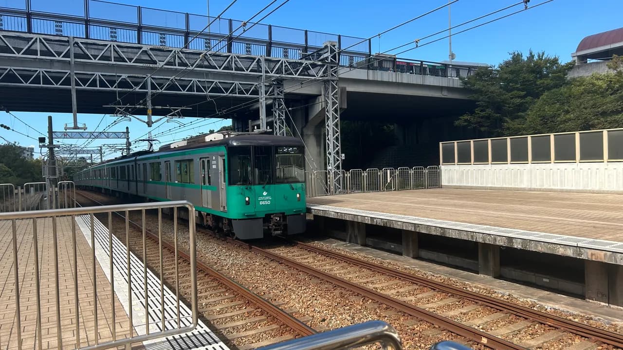 高架道路の下にある地上駅のホームに、緑色の前面の神戸市営地下鉄の電車が停まっている。手前にホームの手すり、上を道路橋が横切り、右に木々が見える