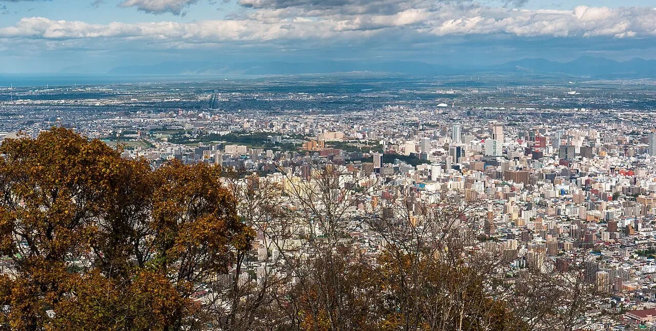 藻岩山から見下ろした昼の札幌市街。手前に色づいた木々、中景にビルが密集する市街地、奥に平野と左奥の海、遠くの山並みまで広がる