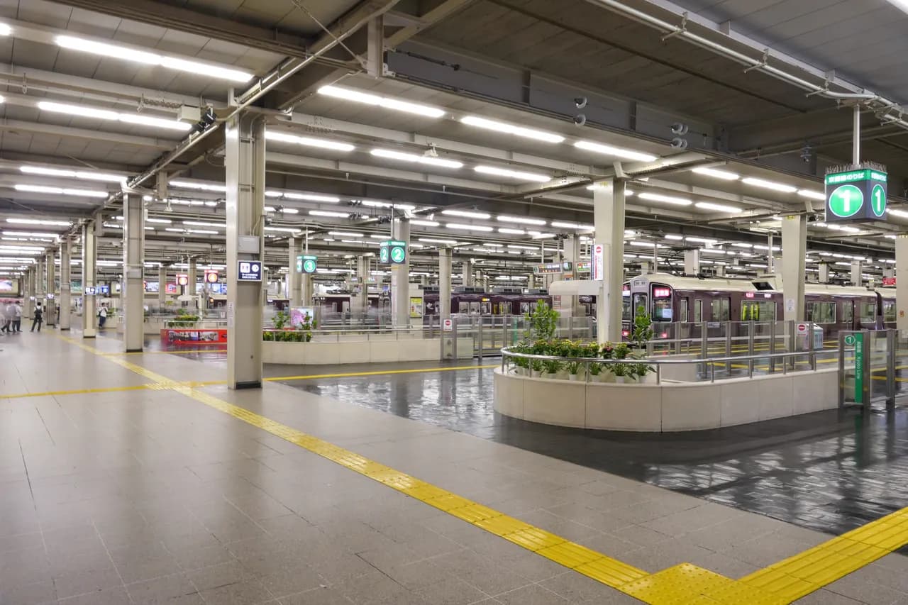 照明が並ぶ広いホーム。床に黄色い点字ブロックが続き、奥の線路に阪急のえんじ色の電車が何本も停まっている
