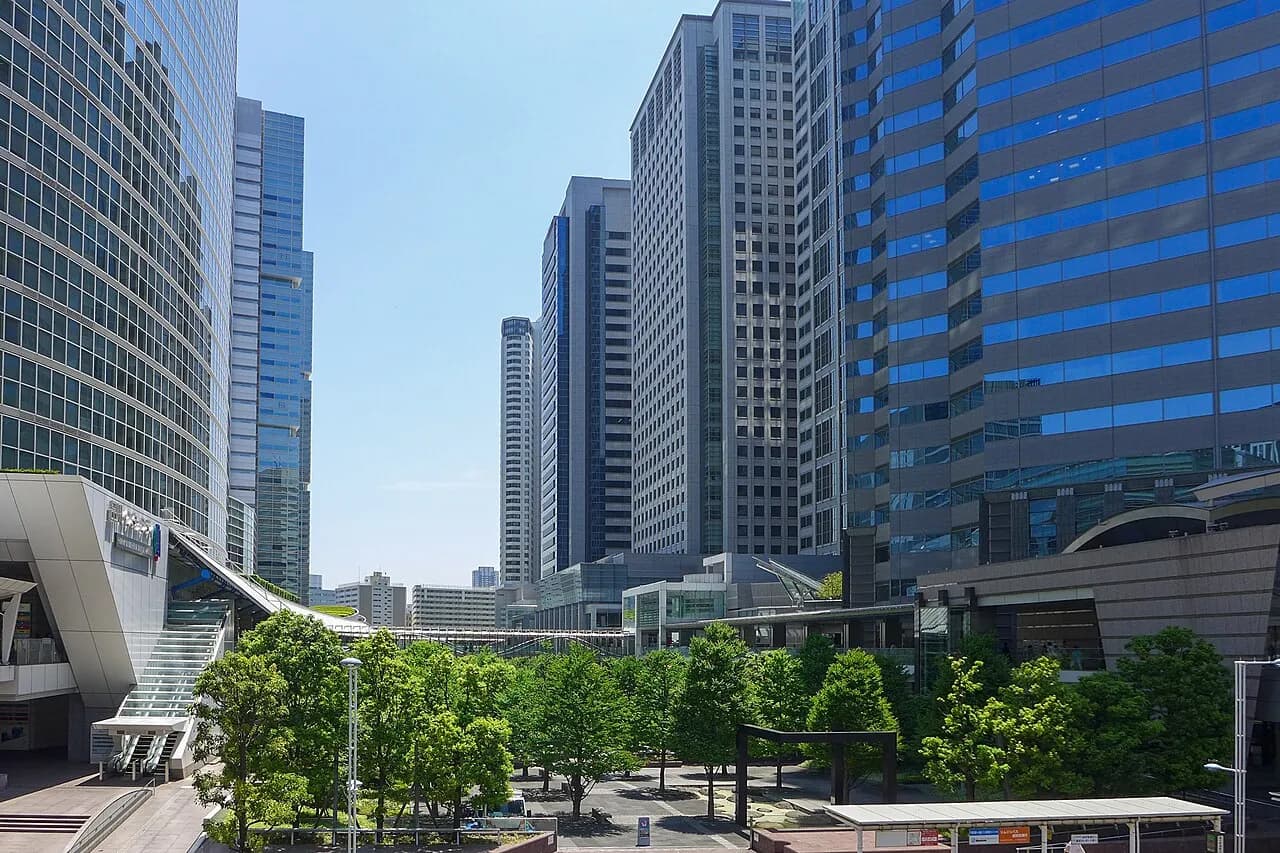 晴天の品川駅港南口側のオフィス街。ガラス張りの高層ビルが並び、手前の広場に緑の木々が茂っている。左手にペデストリアンデッキへ上がる階段が見える
