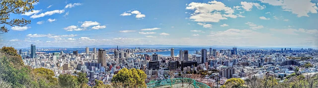 高台から見下ろした晴天の神戸の市街地。高層ビルとマンションが海辺まで続き、その先に神戸港と大阪湾が広がる。手前に木々と緑色の橋の手すりが見える