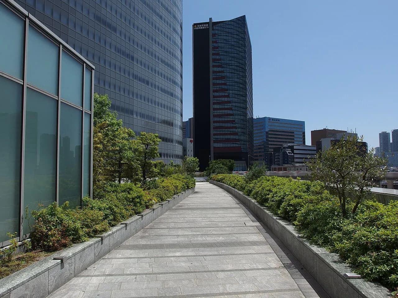 晴天の汐留の高層オフィス街。植え込みに挟まれた石畳の歩道がまっすぐ延び、左手にガラス張りの曲面の高層ビル、正面奥に黒い高層ビルが建つ