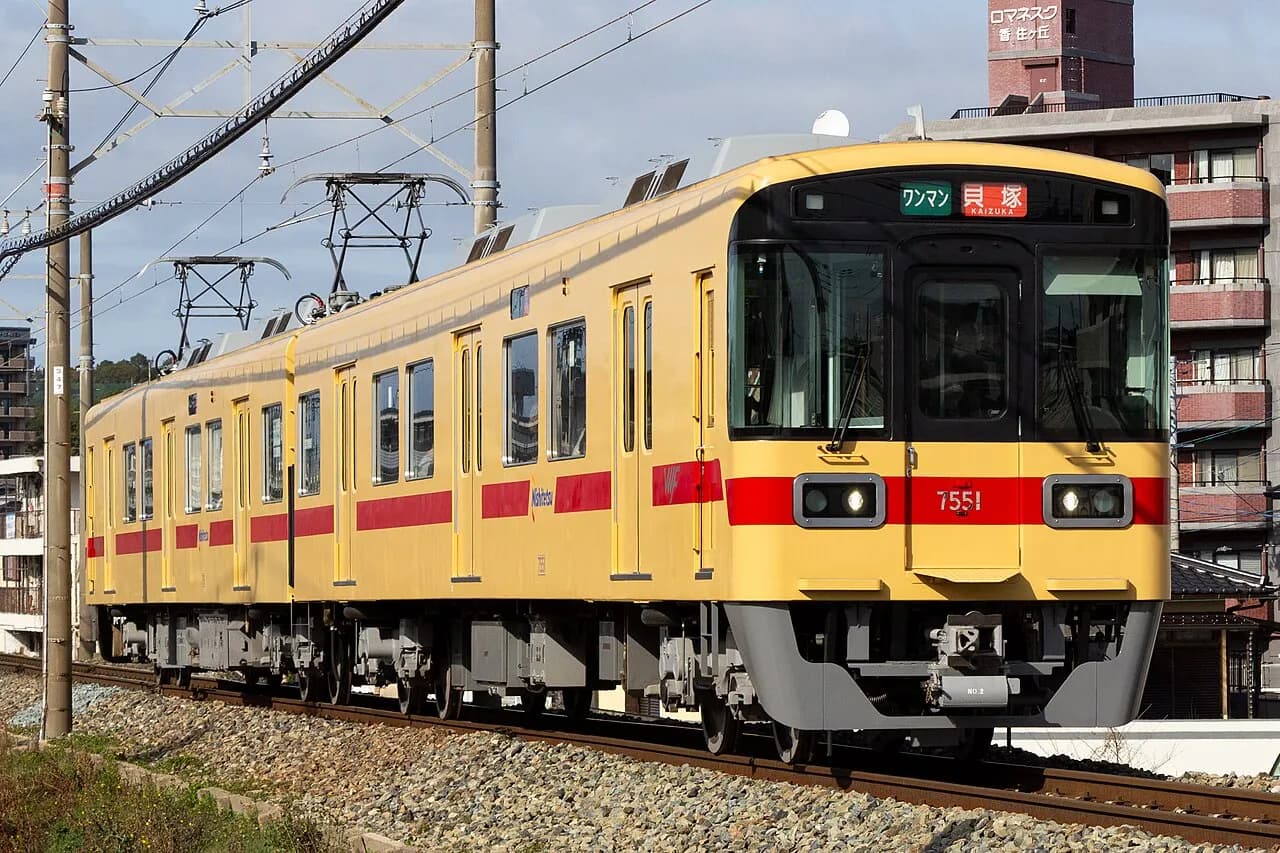 黄色い車体に赤い帯の2両編成の電車が、砂利の線路を走ってくる。行先表示は「貝塚」。背後にれんが色のマンションが建つ