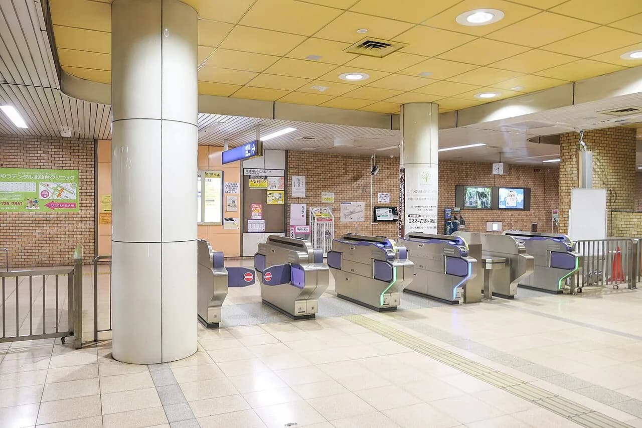 黄色い天井の地下駅の改札口。銀色の自動改札機が6台並び、左右の壁に案内表示や掲示物がある。人はいない