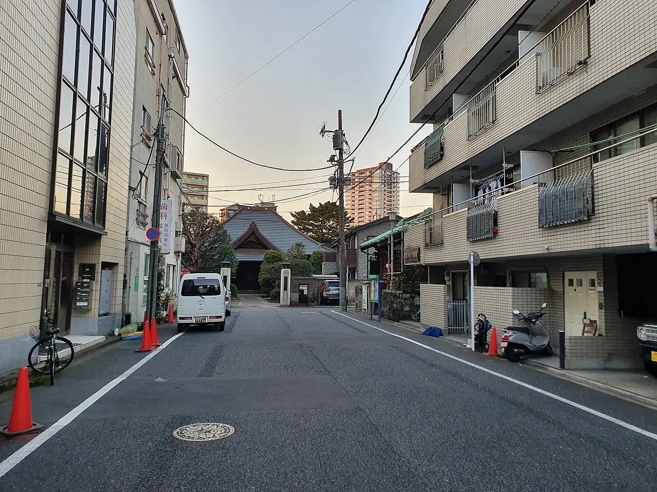 マンションと低層の建物に挟まれた白金の静かな道路。正面奥に瓦屋根の寺の本堂、その向こうに高層マンションが見え、道端に軽自動車のバンとスクーターが止まっている