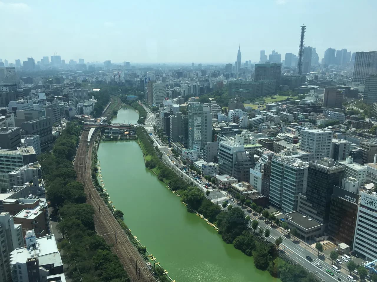 高所から見下ろした外濠。緑色の水面に沿ってJR中央線の線路と並木の外堀通りが延び、両側にオフィスビルが並ぶ。奥に新宿の高層ビル群