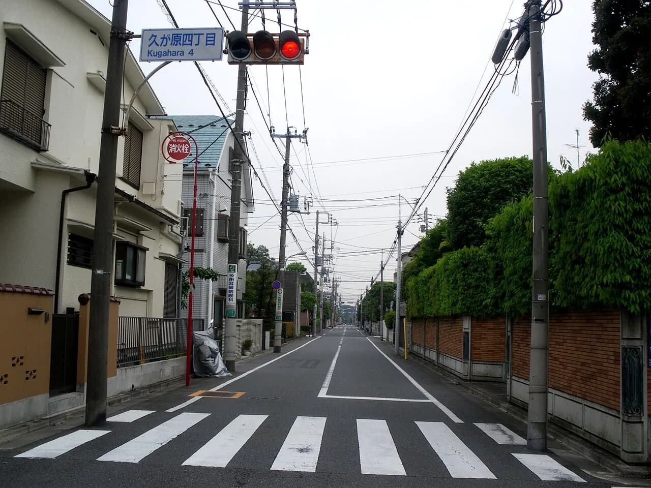 信号機に「久が原四丁目」の地名板が付いた交差点から、まっすぐ延びる片側一車線の道路。両側に戸建て住宅と生け垣が続き、人通りはない