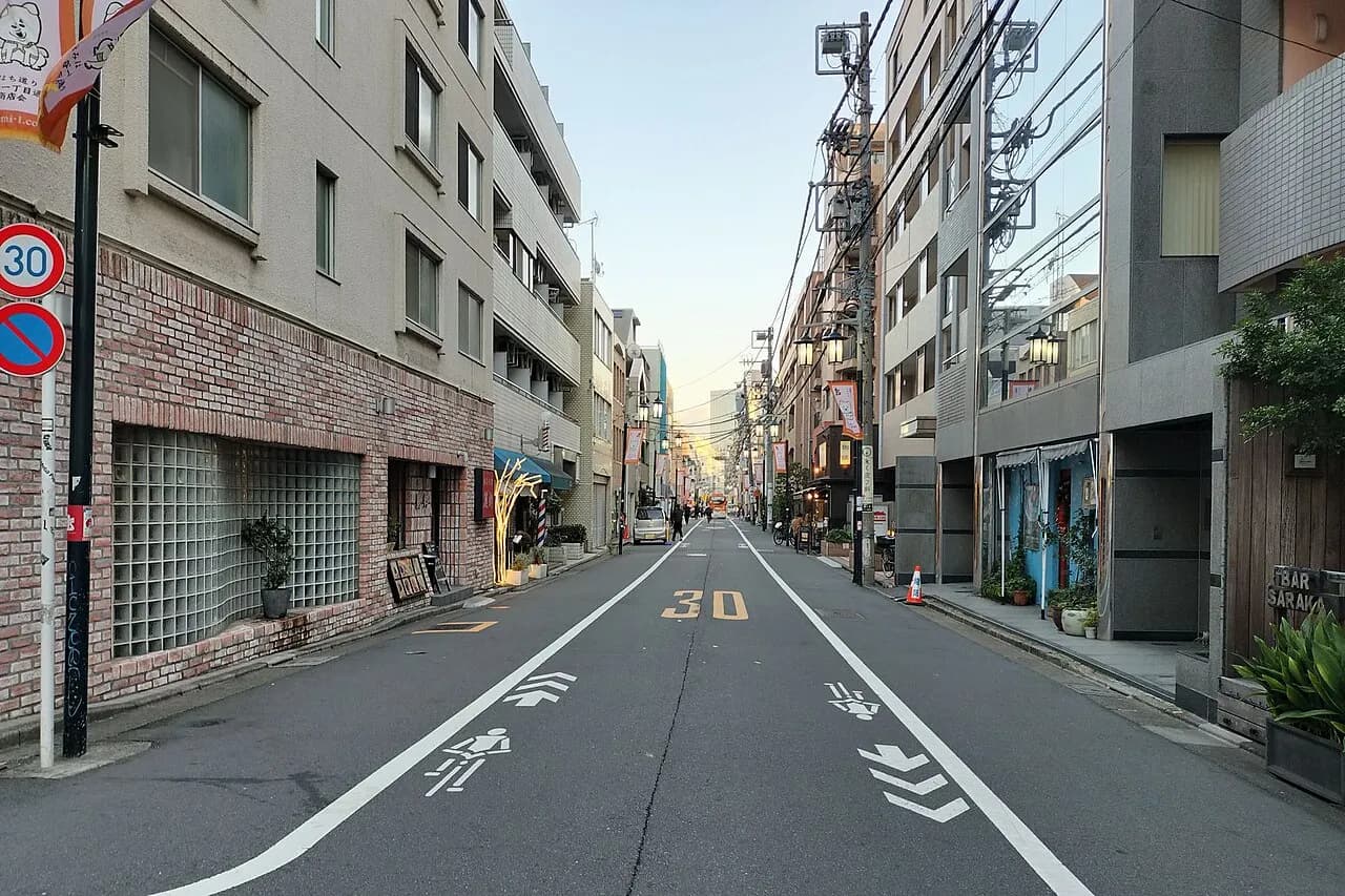 夕方の富ヶ谷の通り。両側に中層のマンションと小さな店が並び、道路には自転車レーンの表示がある。奥へまっすぐ続く道に人影はまばら