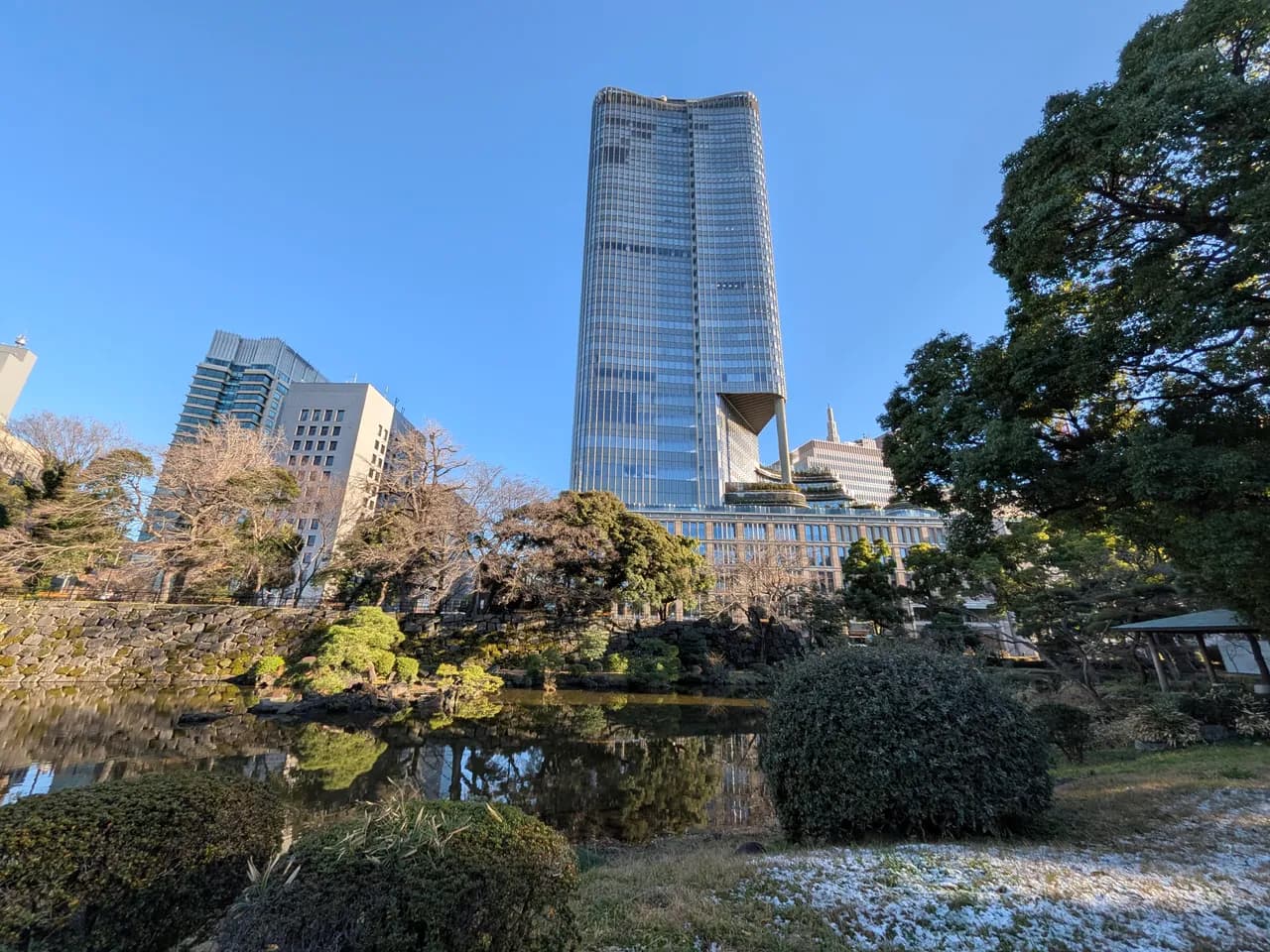 日比谷公園の池と石垣、松などの植え込み越しに見上げたガラス張りの高層ビル。冬の晴天で、手前の芝生にうっすら雪が残っている