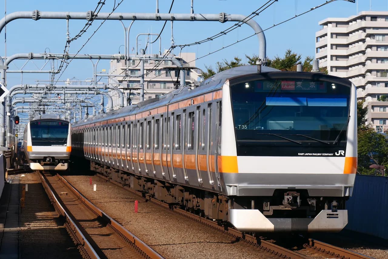 晴れた日の高架線を並んで走る、オレンジ色の帯の中央線快速電車2本。手前の電車の行先表示は「快速 東京」