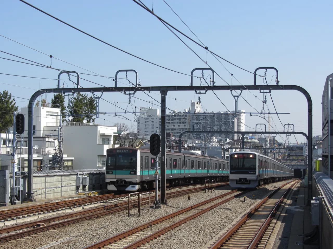 4本並ぶ線路を門形の架線柱がまたぎ、緑の帯のJRの電車と青い帯の小田急の電車が並んでこちらへ向かってくる。背景に住宅とマンションが見える