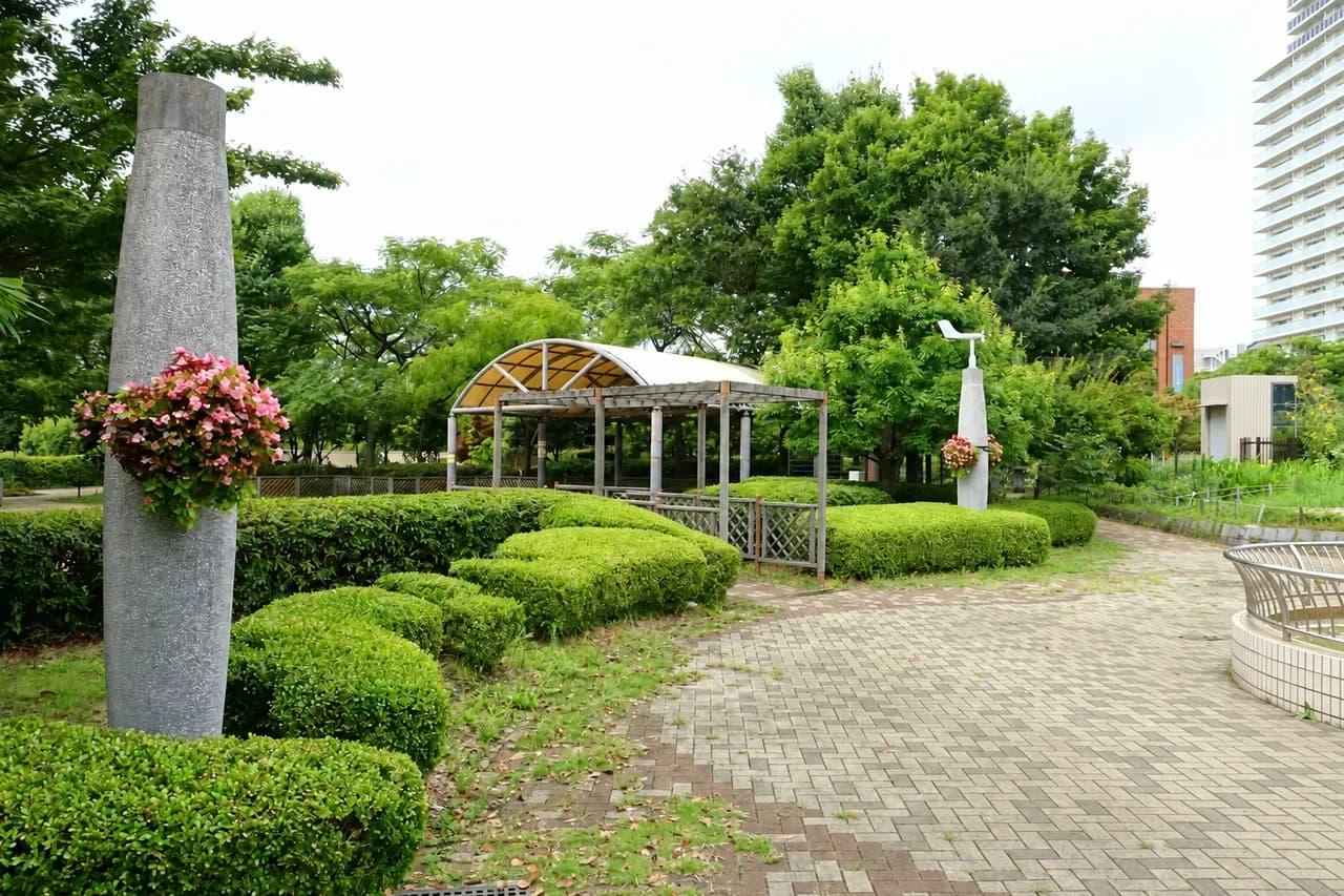 緑の植え込みと藤棚のある木場公園の園路。石柱に花の鉢が飾られ、右奥に高層マンションが見える
