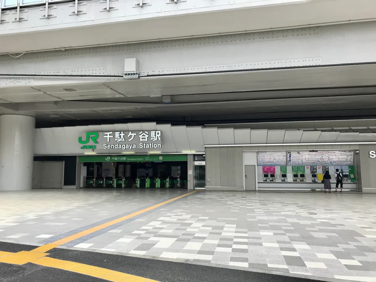 高架の下にある千駄ケ谷駅の改札口。緑の自動改札機が並び、右手の券売機の前に2人が後ろ姿で立つ。手前は石畳の駅前広場